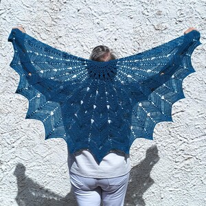 Peut inclure: Un châle fait main, bleu sarcelle foncé, avec un motif ajouré délicat. Le châle est maintenu en l'air, mettant en valeur son motif complexe et dentelé. Les bords du châle ont une forme festonnée et pointue. Fond : mur blanc texturé et ciel bleu clair.