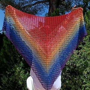 Pode incluir: Um xale de crochê com as cores do arco-íris. O xale transita do vermelho para o laranja, amarelo, verde, azul e roxo. O xale é mostrado em um fundo de folhagem verde e céu azul.