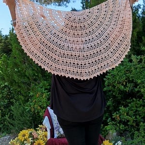 Peut inclure: Un châle en crochet ajouré de couleur pêche clair. Le châle est tenu en l'air, mettant en valeur son design complexe et son bord festonné. L'arrière-plan présente de la verdure et un arbre.