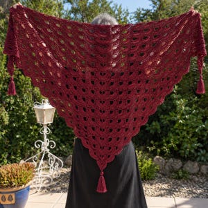 May include: A deep burgundy triangular crochet shawl featuring a repeating fan-like stitch pattern. The edges of the shawl are finished with a scalloped border, and three matching yarn tassels are attached at the two corners and the bottom point.