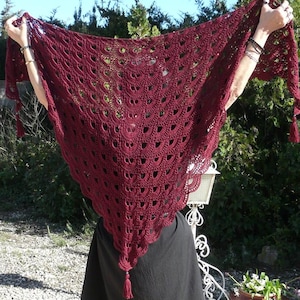 Peut inclure: Un châle en crochet bordeaux avec une bordure festonnée et des glands. Le châle triangulaire est tenu, mettant en valeur le design ajouré complexe. Le châle est de couleur rouge foncé.