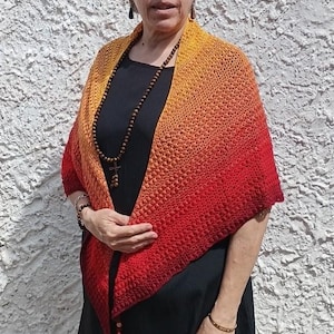 Peut inclure: Une femme porte un châle fait à la main, crocheté. Le châle passe du jaune à l'orange puis au rouge. Elle porte également une robe noire, des lunettes de soleil et un collier de perles en bois.