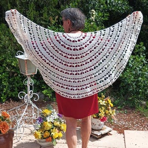 Peut inclure: Un châle en crochet crème à tissage ouvert est présenté, mettant en valeur son design complexe. Le châle est drapé sur une personne portant une robe rouge. L'arrière-plan présente une verdure luxuriante et un cadre extérieur ensoleillé.