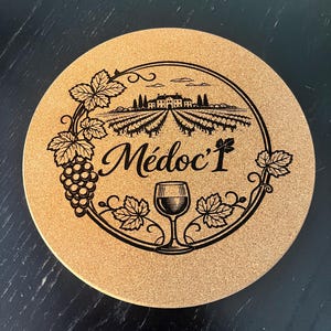 Op de afbeelding: Een ronde onderzetter van kurk met een zwarte lasergegraveerde afbeelding van een wijngaardlandschap met een kasteel, een tros druiven en een wijnglas. De tekst "Médoc" staat centraal in een cirkelvormige rand versierd met wijnbladeren.