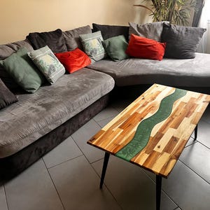 Peut inclure: Une table basse rectangulaire avec un plateau en bois naturel composé de multiples blocs de bois clair et foncé, dotée d'une incrustation centrale en résine verte ondulée et de quatre pieds fins en métal noir, devant un grand canapé d'angle.
