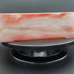 May include: A rectangular resin container with a marbled effect. The colors are primarily white, red, and gold. The container is resting on a black rotating display stand. The resin has a glossy finish.