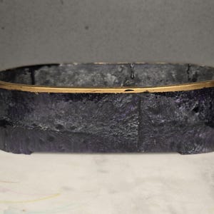 May include: Oval-shaped decorative container with a dark blue and gold rim. The container has a textured surface and a gold-colored rim. The container is sitting on a white surface.
