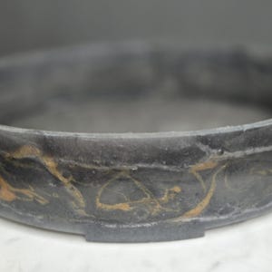 Peut inclure: Un bol décoratif rond gris foncé avec des tourbillons dorés. Le bol a un large bord et une base plate. Le bol est fait d'un matériau marbré et repose sur une surface blanche.