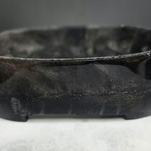 May include: A small, dark gray, rectangular container with rounded corners. The container has a glossy finish and a textured surface. The base has small feet. The container is likely made of a composite material.
