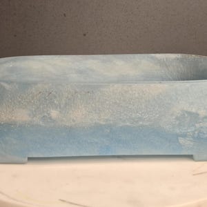 Puede incluir: Una bandeja ovalada de resina con un patrón jaspeado de remolinos azul claro y blancos. La bandeja tiene un acabado liso y brillante con una cualidad translúcida y un borde ligeramente elevado, apoyada sobre una base plana con pequeñas aberturas.