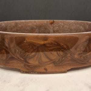 May include: A brown, oval-shaped ceramic planter with a glossy finish. The planter has a decorative swirl pattern and sits on a white surface. The planter is approximately 12 inches long and 6 inches wide.
