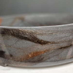 Puede incluir: Una pulsera de resina gris con un diseño veteado que presenta tonos marrones y dorados. La pulsera tiene una forma lisa y redondeada y una calidad sutil y translúcida. Esta pieza única es un accesorio elegante.