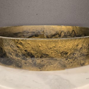 Puede incluir: Un cuenco decorativo redondo y de perfil bajo con un diseño veteado en oro y negro. El cuenco está hecho de un material brillante y se asienta sobre una superficie de mármol blanco. El diámetro del cuenco es de unos 25 cm.