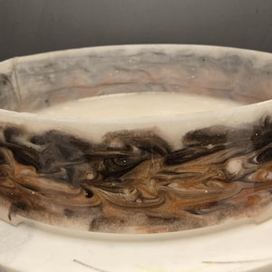 May include: A round, clear resin bowl with a marbled design. The bowl features swirls of brown, black, and gold colours. The interior of the bowl is a creamy white colour. The bowl is sitting on a white marble surface.