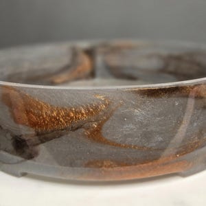 Puede incluir: Un cuenco redondo de resina gris con un diseño de remolino metálico bronce y negro. El cuenco tiene un acabado liso y brillante y se asienta sobre una superficie blanca. El interior del cuenco presenta un efecto veteado.