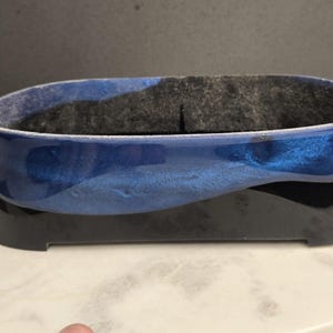 May include: A blue and black decorative container with an oval shape. The container has a glossy finish, with a dark blue top and a black base. The interior is lined with a dark gray material. The container sits on a white surface.