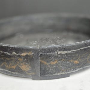 May include: A dark gray, circular object with a band of gold-colored decorative elements. The object appears to be made of a stone-like material. The object is resting on a white surface.