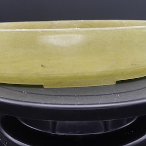 May include: A light green, oval-shaped container with a flat base. The container is made of a translucent material and sits on a black, circular platform. The container's walls are smooth and the overall design is simple.