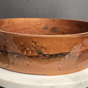 Könnte beinhalten: Eine kupferfarbene, runde Schüssel mit niedrigem Rand und strukturierter Oberfläche. Die Schüssel hat ein dunkles Band in der Mitte und steht auf einer weißen Marmoroberfläche. Die Außenseite der Schüssel hat eine raue, unebene Textur.
