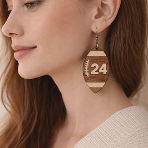 May include: Wooden football-shaped earrings with the number 24 in white. The earrings have a dark brown wood grain with two horizontal white stripes. The earrings are hanging from a gold-colored hook.
