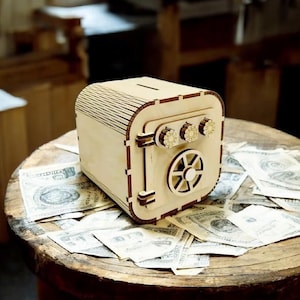 May include: A wooden bank shaped like a safe, with a slot on top and a door with a dial. The bank is light brown and sits on a wooden table covered in dollar bills. The safe has three gold-colored dials.