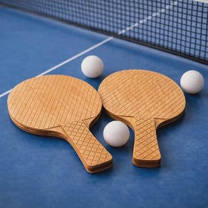 May include: Two wooden ping pong paddles with a textured surface and three white ping pong balls rest on a blue table. A net is visible in the background, suggesting a game of table tennis.