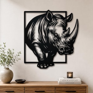 May include: Black metal wall art of a rhinoceros. The rhino is depicted in a detailed, stylized illustration, partially framed by a black square. The artwork is mounted on a white wall, creating a striking contrast. The rhino's horn and features are clearly defined.