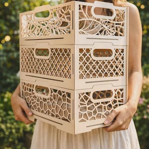 Puede incluir: Tres cajas de madera apiladas, de color claro, con patrones decorativos recortados. Cada caja tiene una abertura rectangular para el asa. Las cajas son sostenidas por una persona que lleva un sombrero de paja y un vestido color crema.