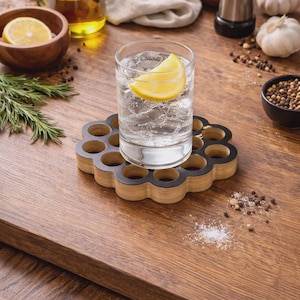 Puede incluir: Un vaso de agua con gas y una rodaja de limón descansa sobre un posavasos de madera con detalles negros. El posavasos está rodeado de ingredientes como ajo, granos de pimienta y romero.