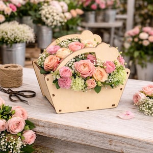 Può includere: Un cestino di legno pieno di rose rosa e pesca, garofani e gipsofila. Il cestino ha un manico con intagli a forma di cuore. La composizione è su un tavolo di legno invecchiato, con altre composizioni floreali sullo sfondo.