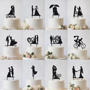 Puede incluir: Una colección de adornos para tartas con siluetas negras que representan parejas en varias poses. Algunos adornos incluyen texto como "Mr & Mrs" y "Love". Los adornos se muestran en pasteles glaseados blancos decorados con flores, perfectos para bodas.