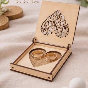 May include: Laser cut wooden wedding rings box. The box is light brown with a heart-shaped cutout on the lid. Inside, two gold rings rest on a burlap heart. The box measures 12 x 12 x 1.7 cm.
