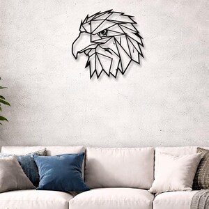 May include: A black metal wall art piece depicting an eagle's head in a geometric, polygonal design. The artwork is mounted on a textured white wall above a white sofa with blue and gray throw pillows.