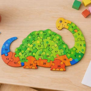 May include: A wooden dinosaur puzzle with 29 pieces, featuring green, orange, and blue sections. The puzzle is on a light wooden surface, with colored pencils and a drawing of a dinosaur nearby. The puzzle is numbered.