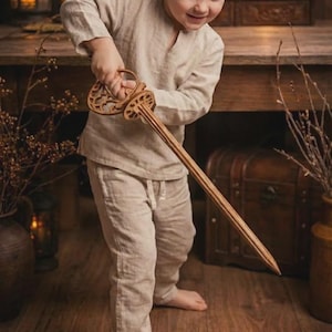 May include: A young child holds a wooden toy sword with an ornate handle. The sword is light brown and appears to be made of wood. The child is wearing a light-colored linen outfit. The background includes wooden furniture and decorative items.