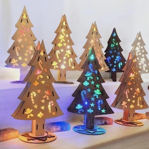 May include: A selection of wooden Christmas tree decorations with cut-out designs. The trees are lit with LED lights, displaying candy canes, stars, and gingerbread men. The trees are brown, with some painted dark brown or green. The bases are various colors.