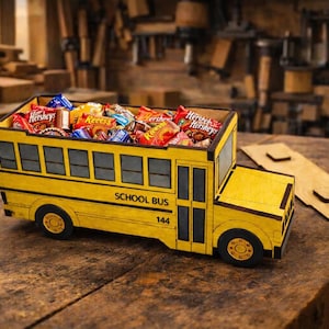 May include: A yellow wooden school bus-shaped container filled with assorted candies. The bus features black wheels, windows, and the words "SCHOOL BUS" and the number "144". The container sits on a wooden surface.