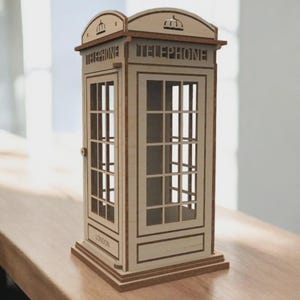 Könnte beinhalten: Ein Miniatur-Telefonzelle aus Holz mit dem Wort "TELEPHONE" auf der Oberseite und den Seiten. Die Zelle hat einen hellbraunen Rahmen und klare Fenster. Das Wort "LONDON" ist eingraviert.