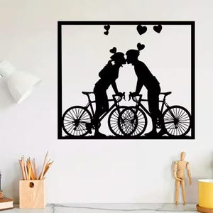 Könnte beinhalten: Schwarze Silhouette eines Kunstwerks, das ein sich küssendes Paar über Fahrrädern mit Herzakzenten zeigt. Das Kunstwerk ist in einem schwarzen Quadrat gerahmt, wobei die Köpfe des Paares mit Herzformen verziert sind. Die Fahrräder sind mit Speichen und Lenkern detailliert.