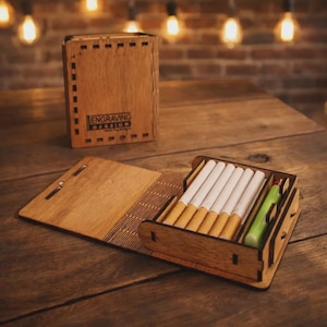 May include: Wooden cigarette case with a hinged lid, holding several cigarettes. The case is open, revealing the cigarettes and a green lighter. A matching wooden box with the text "ENGRAVING DESIGN" is in the background.
