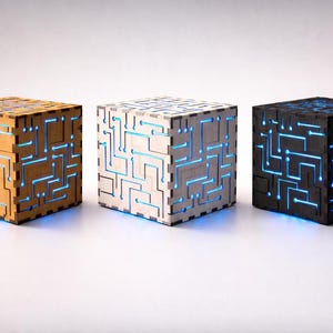 Puede incluir: Tres lámparas cúbicas con diseño de placa de circuito, en tonos beige, blanco y negro. Cada lámpara tiene luces LED azules. Las lámparas son de madera y tienen una estética moderna e inspirada en la tecnología.