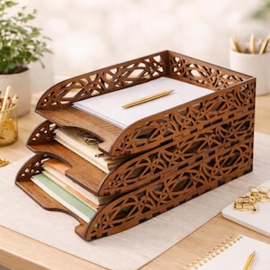 Può includere: Un organizer da scrivania in legno a tre livelli con un design decorativo traforato. L'organizer contiene pile di carta e una penna dorata. Il legno ha un caldo tono marrone e l'organizer è su una scrivania chiara.