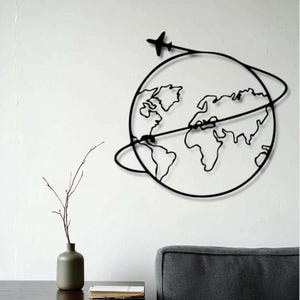 May include: Black metal wall art showing a globe with continents. A small aeroplane flies around the globe. The artwork is mounted on a white wall. A vase and a book are visible below. The piece is ideal for travel enthusiasts.