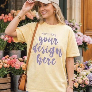 May include: A light yellow t-shirt with the text "your design here" in purple script. The shirt is worn with a beige baseball cap and denim shorts. A brown shoulder bag is also visible.