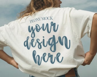 Op de afbeelding: Wit T-shirt met de tekst "your design here" in een blauw scriptlettertype. Het shirt maakt deel uit van een mock-up, met de woorden "Peony Mock" boven de hoofdtekst. Het model draagt een roze pet en een blauwe jeans.