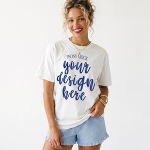 Puede incluir: Camiseta blanca con el texto "PEONY MOCK" y "your design here" en escritura azul. La modelo lleva pantalones cortos azul claro con dobladillo con volantes. La modelo tiene el pelo rubio y lleva joyas de oro.