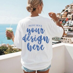 Op de afbeelding: Wit T-shirt met de tekst "your design here" in blauw script. Het shirt is een mock-up, ideaal om ontwerpen te presenteren. Het model draagt een spijkerbroekshort en houdt een groene drank met een rietje vast. De achtergrond toont een kustlandschap.