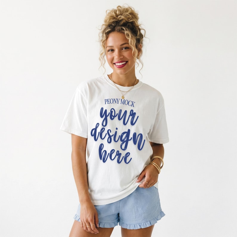 Puede incluir: Camiseta blanca con el texto "PEONY MOCK" y "your design here" en escritura azul. La modelo lleva pantalones cortos azul claro con dobladillo con volantes. La modelo tiene el pelo rubio y lleva joyas de oro.