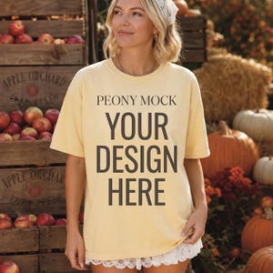 May include: A pale yellow short-sleeved t-shirt featuring the dark brown text PEONY MOCK YOUR DESIGN HERE printed on the front. The shirt has a crew neckline and a straight hemline with a white lace trim detail along the bottom edge.