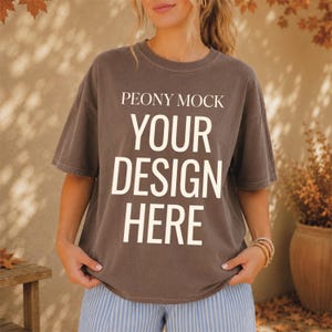 以下が含まれることがあります： 胸元に太字のサンセリフ体で白いテキスト「PEONY MOCK YOUR DESIGN HERE」がプリントされた、ダークブラウンの半袖Tシャツ。クルーネックでゆったりとしたフィット感があり、水色のストライプ柄のパンツと組み合わされています。